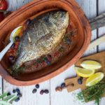 Gebackener Fisch in einer Terrakottaform mit Tomaten und Oliven, daneben Zitronenscheiben und frische Tomaten.
