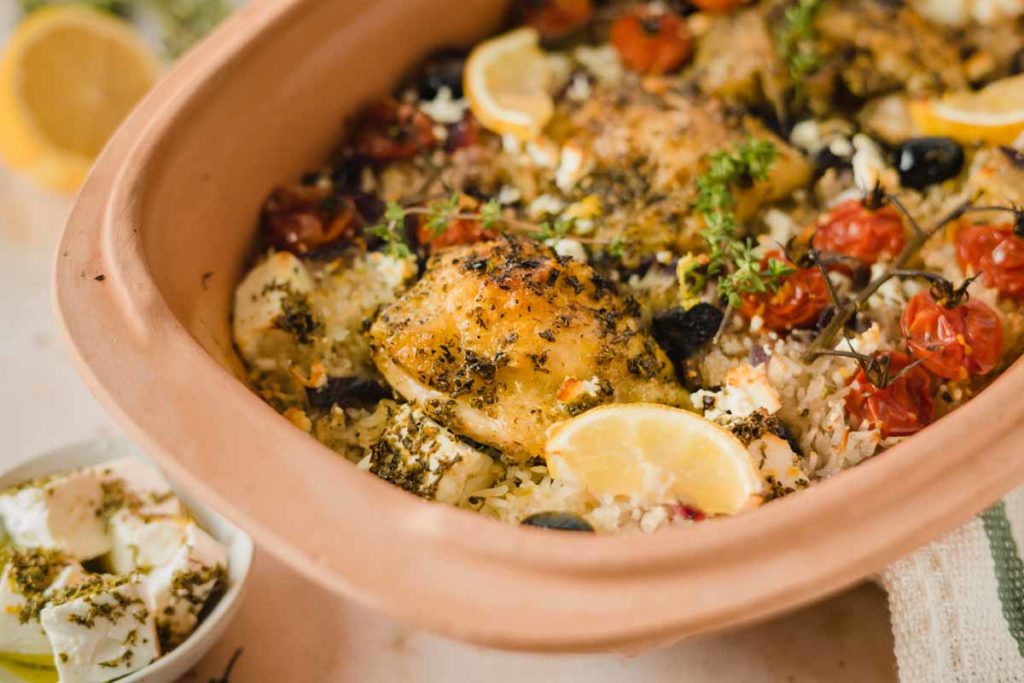 Mediterranes Ofengericht mit Hühnchen, Reis, Kirschtomaten, Feta und Zitronenscheiben in einer Keramikform.