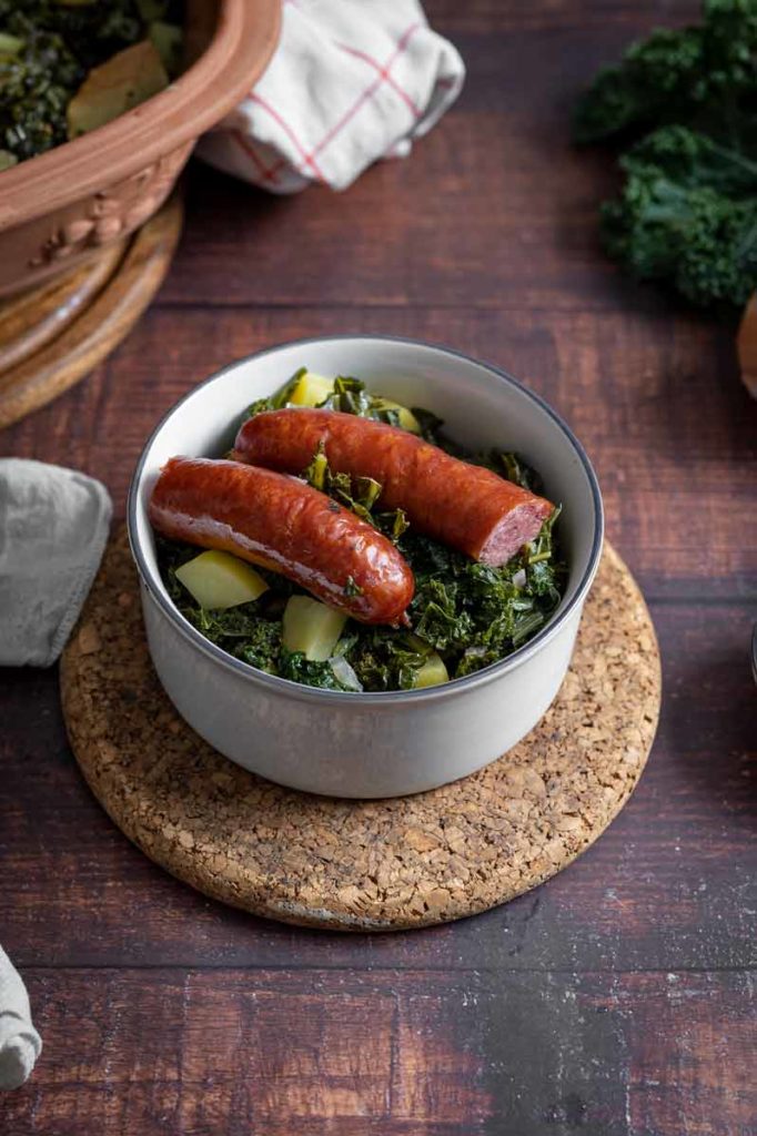 Schüssel mit Grünkohl, Kartoffeln und zwei Mettwürsten auf einem Korkuntersetzer auf Holztisch.