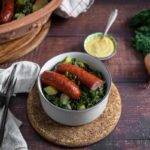 Schüssel mit Grünkohl, Kartoffeln und zwei Würsten auf einem Tisch, daneben Senf, Zwiebel und Grünkohlblätter.