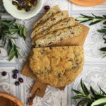 Olivenbrot auf Holzbrett, geschnitten mit einer Schüssel Oliven daneben, umgeben von Olivenzweigen.