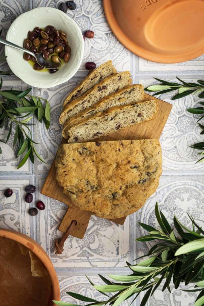 Olivenbrot auf Holzbrett, geschnitten mit einer Schüssel Oliven daneben, umgeben von Olivenzweigen.