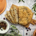 Geschnittenes Olivenbrot auf Holzbrett mit Oliven in Schale und zwei Terrakotta-Gärschalen, dekoriert mit Olivenzweigen.