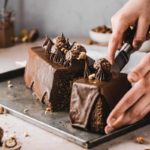 Hände schneiden einen Schokoladenkuchen mit Pralinen und Schokosahne auf Blech an.