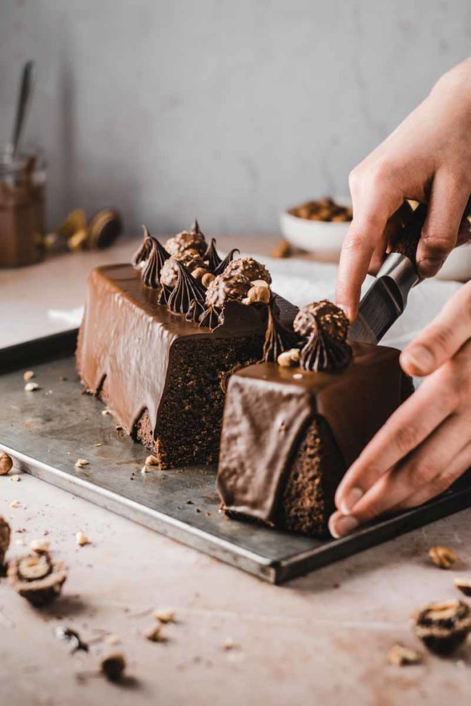 Hände schneiden einen Schokoladenkuchen mit Pralinen und Schokosahne auf Blech an.