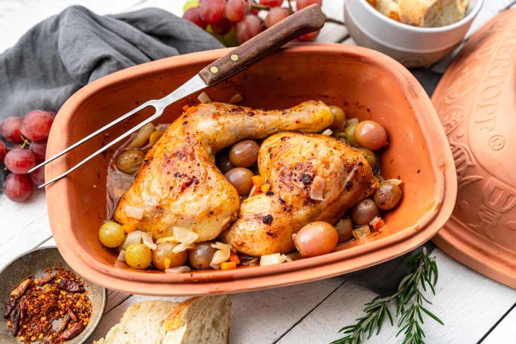 Gebratenes Hähnchen mit Trauben und Gemüse im Bräter, umgeben von Brot, Gewürzen und frischen Trauben.