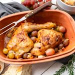 Gebratenes Hähnchen mit Trauben und Gemüse im Bräter, umgeben von Brot, Gewürzen und frischen Trauben.