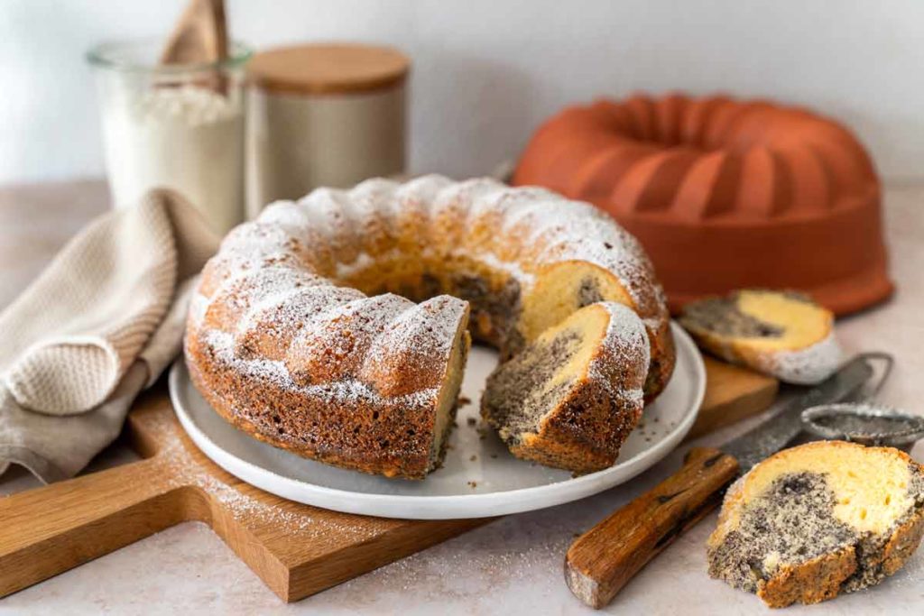 Marmor-Gugelhupf mit Puderzucker bestäubt auf weißem Teller, angeschnitten, daneben Backform und Zutaten.