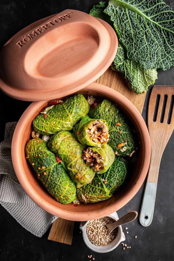 Wirsingrouladen gefüllt mit Reis und Gemüse in einem Römertopf auf schwarzem Untergrund, daneben frische Wirsingblätter.