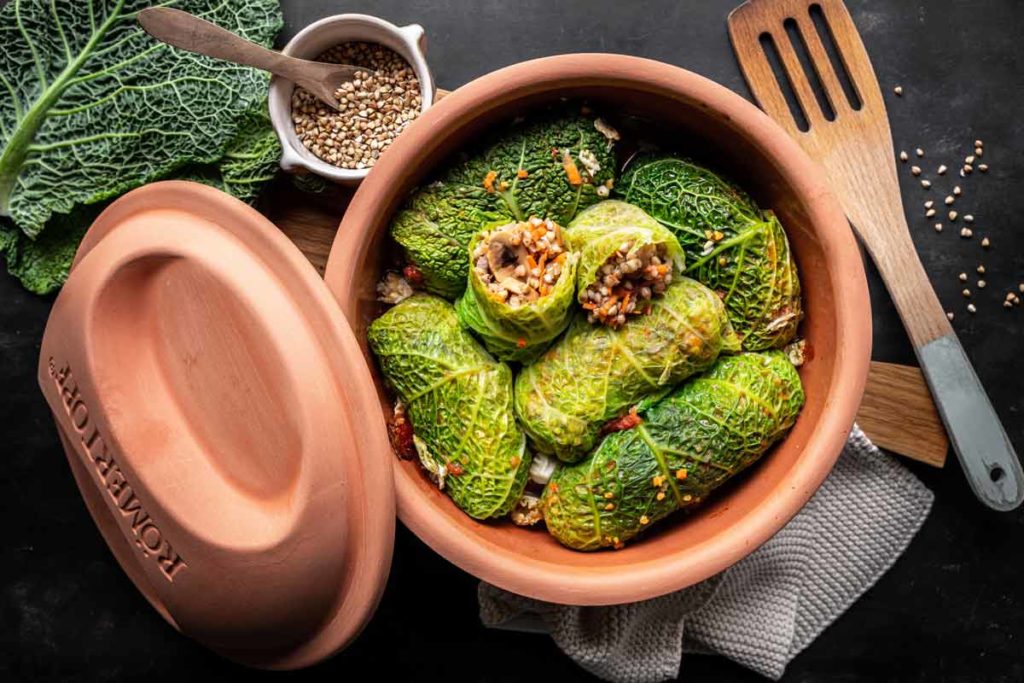 Kohlrouladen im Tontopf, gefüllt mit Gemüse und Getreide, mit einem Holzlöffel und frischem Wirsing daneben.