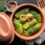 Kohlrouladen im Tontopf, gefüllt mit Gemüse und Getreide, mit einem Holzlöffel und frischem Wirsing daneben.