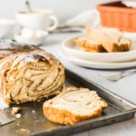 Geschnittenes geflochtenes Zimtgebäck mit Zuckerguss auf Backblech, im Hintergrund Tasse und weitere Scheiben auf Tellern.