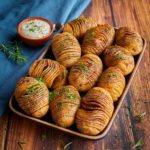 Gebackene Fächerkartoffeln mit Kräutern auf einem Tablett, daneben eine Schale Dip auf einem Holztisch.