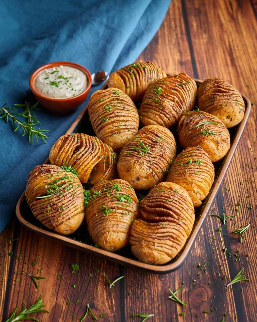 Gebackene Fächerkartoffeln mit Kräutern auf einem Tablett, daneben eine Schale Dip auf einem Holztisch.