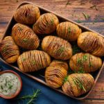 Hasselback-Kartoffeln auf einem Blech mit Kräutern, daneben eine Schale mit Dip, auf Holztisch angerichtet.