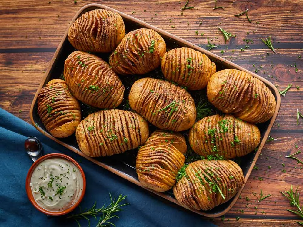 Hasselback-Kartoffeln auf einem Blech mit Kräutern, daneben eine Schale mit Dip, auf Holztisch angerichtet.