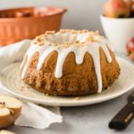 Gugelhupf mit weißer Glasur auf Teller, Apfelhälfte und Messer daneben, im Hintergrund Äpfel und Backform.