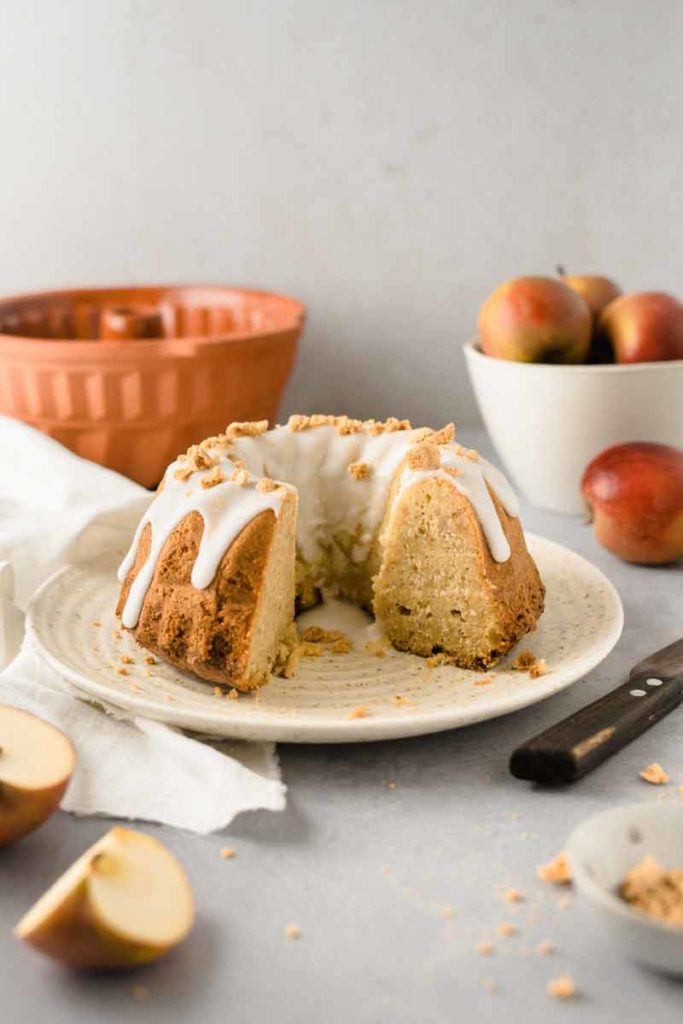Gugelhupf mit Zuckerguss und Krümeln auf einem Teller, daneben frische Äpfel und eine Gugelhupfform im Hintergrund.