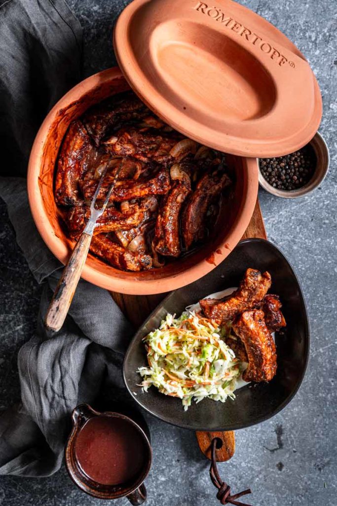 Römertopf mit marinierten Rippchen, serviert mit Krautsalat und Soße, auf einem rustikalen Tisch gedeckt.