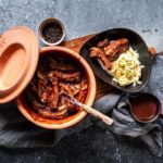 Tontopf mit marinierten Rippchen, Servierteller mit Rippchen und Krautsalat, Schale mit Pfefferkörnern auf grauem Untergrund.