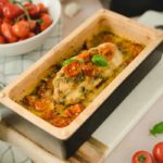 Gebackenes Hähnchenfilet mit Tomaten und Kräutern in einer Auflaufform. Hintergrund: Kirschtomaten und Pesto.