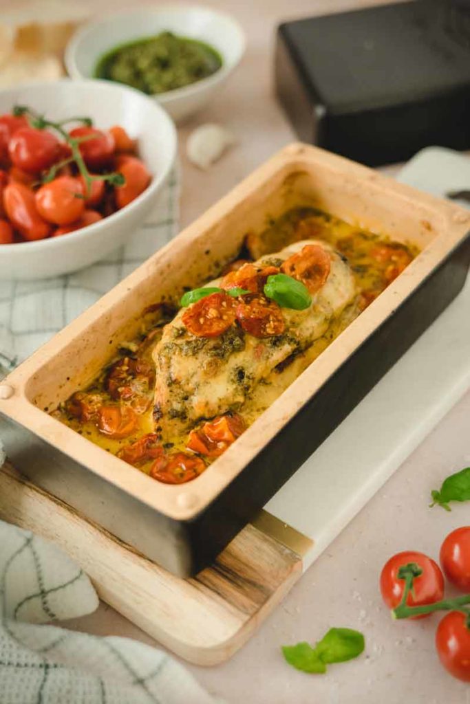 Gebackenes Hähnchenfilet mit Tomaten und Kräutern in einer Auflaufform. Hintergrund: Kirschtomaten und Pesto.