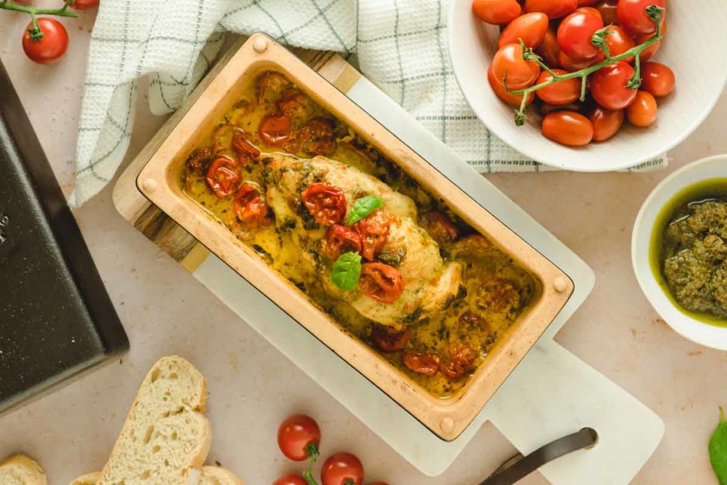 Gebratenes Hähnchenbrustfilet mit Tomaten und Kräutern in einer Auflaufform, umgeben von frischen Tomaten und Brot.