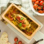 Gebratenes Hähnchenbrustfilet mit Tomaten und Kräutern in einer Auflaufform, umgeben von frischen Tomaten und Brot.
