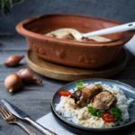 Ein Teller mit Reis, Fleisch, Tomaten und Petersilie. Im Hintergrund ein Tontopf mit Schöpfkelle.