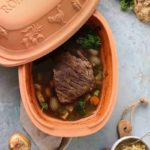 Römertopf mit Braten und Gemüse auf grauem Untergrund, umgeben von Zwiebel, Sellerie und Karotte.