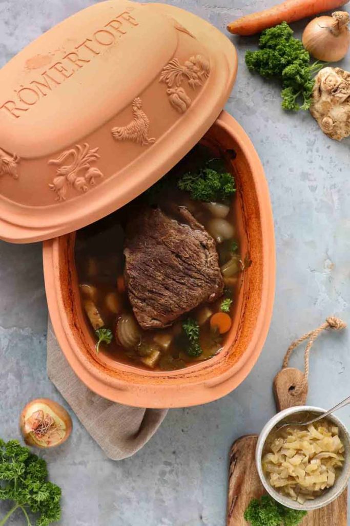 Römertopf mit Braten und Gemüse auf grauem Untergrund, umgeben von Zwiebel, Sellerie und Karotte.