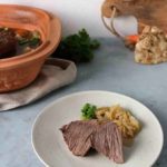 Gekochtes Rindfleisch mit Zwiebeln auf einem Teller, im Hintergrund ein Römertopf und Gemüse, auf einem grauen Tisch arrangiert.