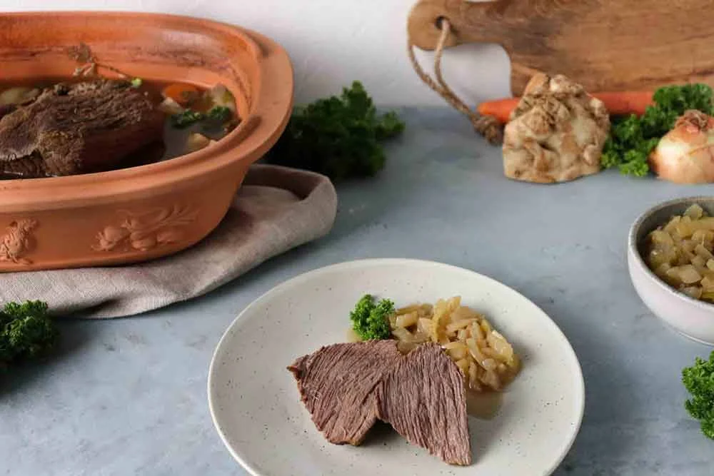 Gekochtes Rindfleisch mit Zwiebeln auf einem Teller, im Hintergrund ein Römertopf und Gemüse, auf einem grauen Tisch arrangiert.