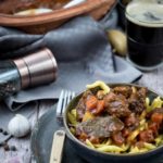 Schüssel mit Rindergulasch und Spätzle, daneben ein Glas dunkles Bier, Pfeffermühle und Knoblauch auf Holztisch.