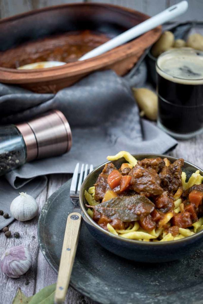 Schüssel mit Rindergulasch und Spätzle, daneben ein Glas dunkles Bier, Pfeffermühle und Knoblauch auf Holztisch.