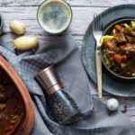 Serviertes Gulasch mit Spätzle auf einem Teller, daneben Kartoffeln, Pfeffermühle, Knoblauch und eine Schüssel mit Löffel.