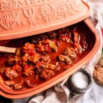 Gulasch in einem Römertopf mit Holzlöffel, dazu eine Zwiebel, Brot und ein roter Paprika auf einem weißen Tisch.