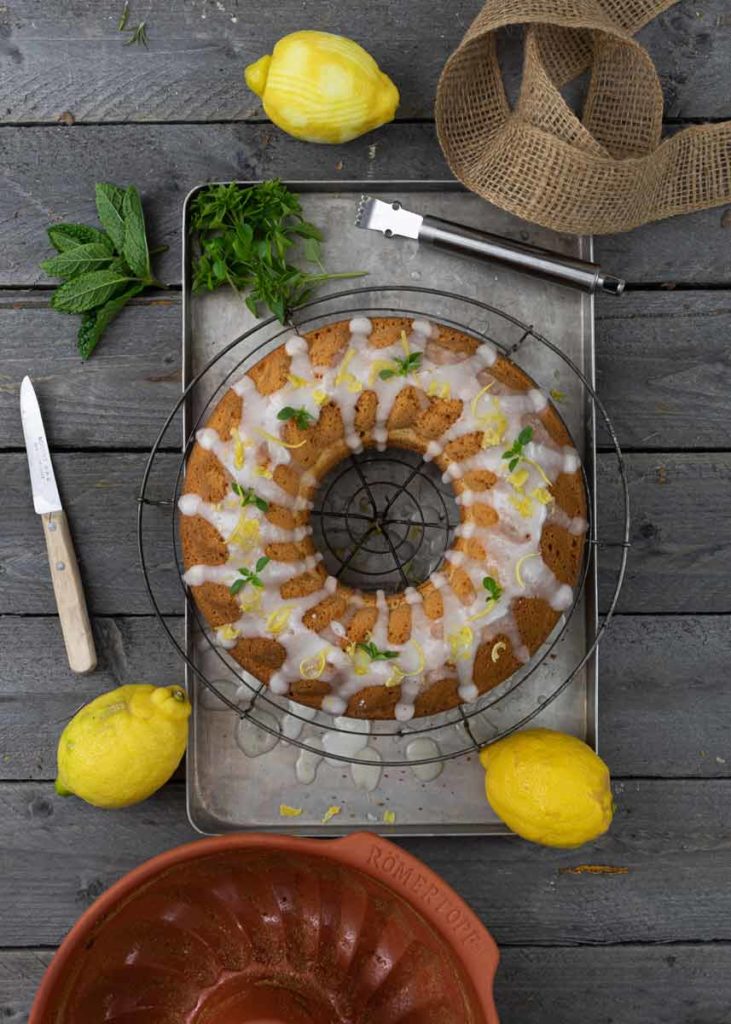Zitronenkuchen mit Zuckerglasur auf einem Gitterrost, umgeben von Zitronen, Minze und einem Messer auf Holztisch.