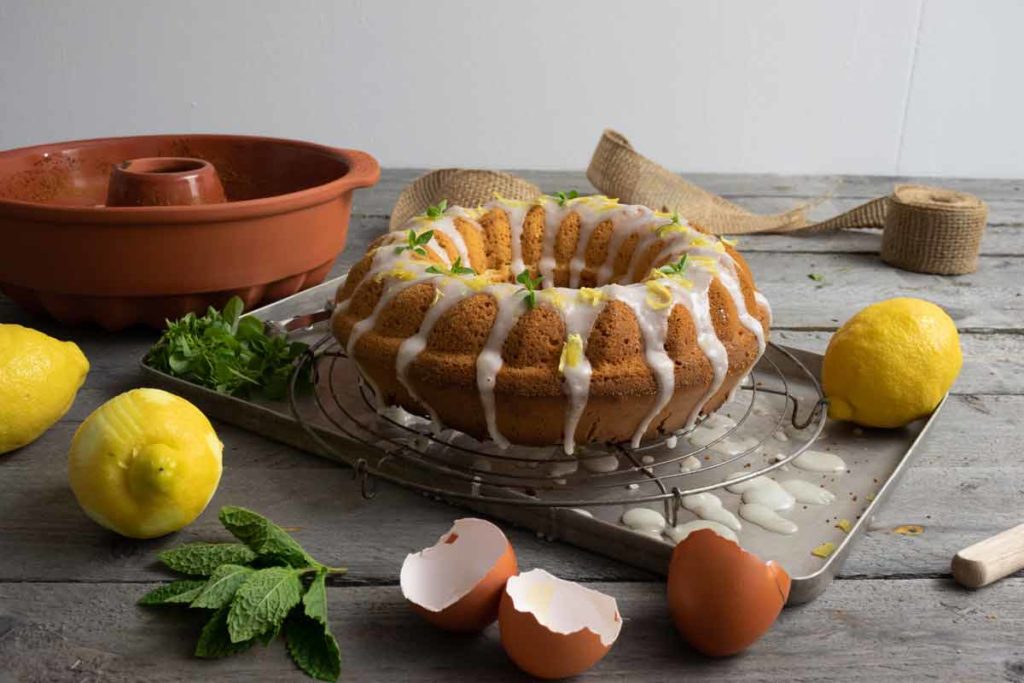 Zitronenkuchen mit Zuckerglasur auf einem Gitter, daneben Zitronen, Eierschalen und frische Kräuter auf einem Holztisch.