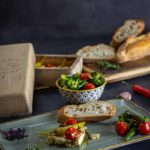 Baguette mit gebackenem Feta, Tomaten, Peperoni und Brokkoli auf einem Teller mit Brot und Gemüse im Hintergrund.