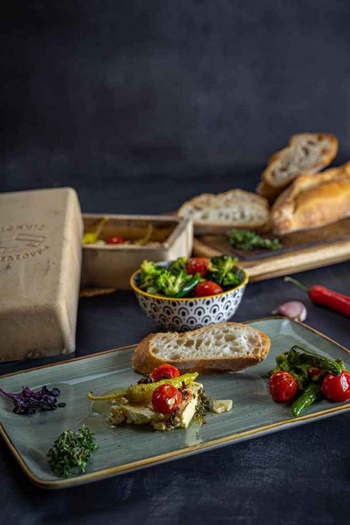 Baguette mit gebackenem Feta, Tomaten, Peperoni und Brokkoli auf einem Teller mit Brot und Gemüse im Hintergrund.