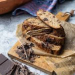 Ein geschnittenes Bananenbrot mit Schokoladenstücken auf Holzbrett, daneben blaue Serviette.