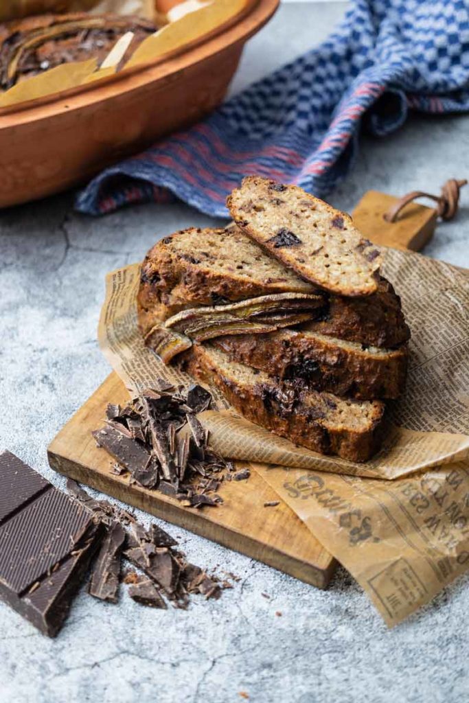 Ein geschnittenes Bananenbrot mit Schokoladenstücken auf Holzbrett, daneben blaue Serviette.
