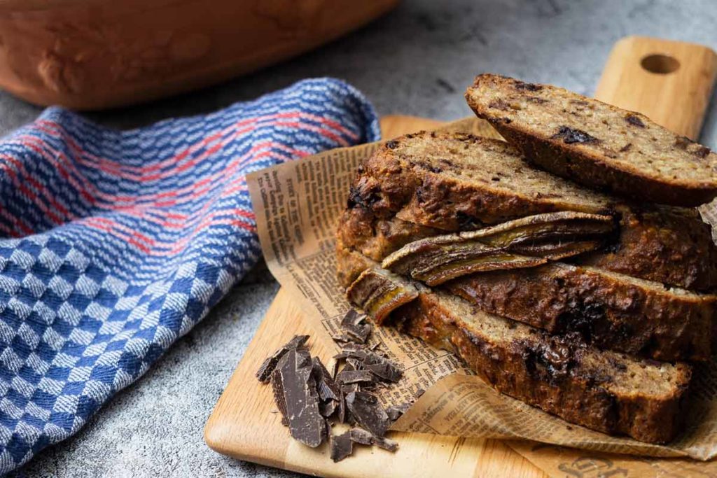 Geschnittenes Bananenbrot mit Schokostücken auf Holzbrett, daneben blau-rot-kariertes Tuch.