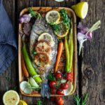 Gebackener Fisch mit Zitronenscheiben, buntem Gemüse und Kräutern auf Holzuntergrund.