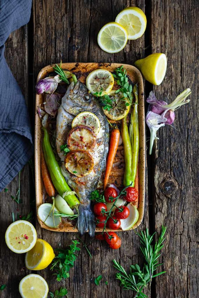 Gebackener Fisch mit Zitronenscheiben, buntem Gemüse und Kräutern auf Holzuntergrund.