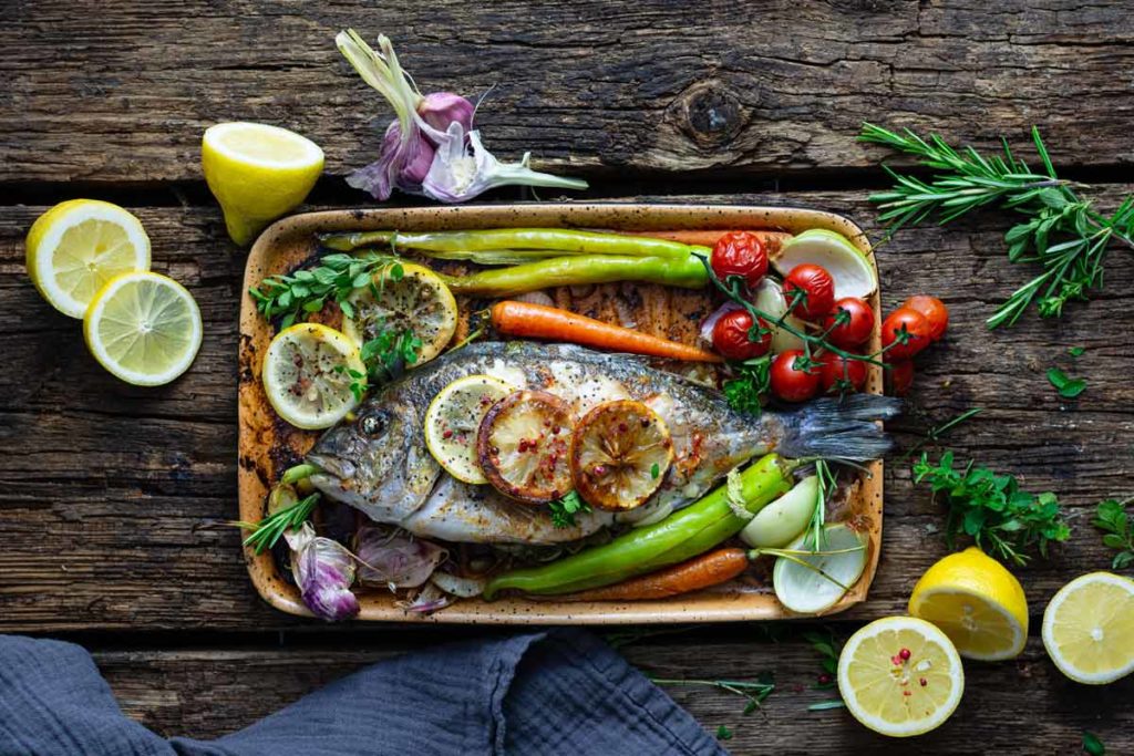 Gebackener Fisch mit Zitronenscheiben, Tomaten, Paprika und Kräutern auf einem Holzbrett, umgeben von frischen Zutaten.