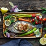 Gebackener Fisch mit Zitronenscheiben, Tomaten, Paprika und Kräutern auf einem Holzbrett, umgeben von frischen Zutaten.