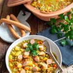 Schüssel mit Erbseneintopf, gewürfelten Karotten, Kartoffeln und geschnittenen Würstchen, daneben Baguette und ein Römertopf.