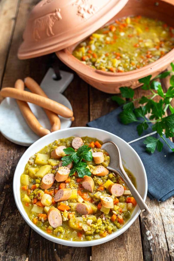 Schüssel mit Erbseneintopf, gewürfelten Karotten, Kartoffeln und geschnittenen Würstchen, daneben Baguette und ein Römertopf.